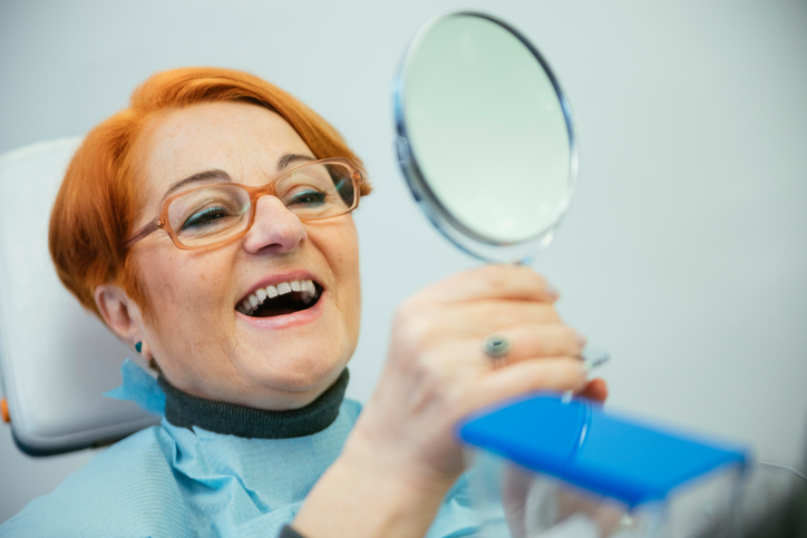 Senior Woman Sitting In A Chair Looking At Her Teeth In The Mirror
